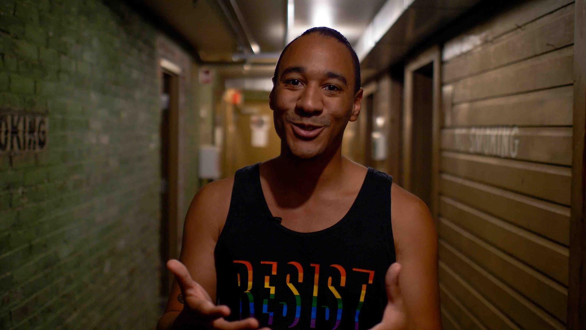 Caleb stands in a bright hallway with his arms out towards the camera. His palm are open as if he is greeting us. He is wearing a black tanktop with the word 'Resist' which is also in black but its form made clear thruough the use of a rainbow dropshadow on each letter.